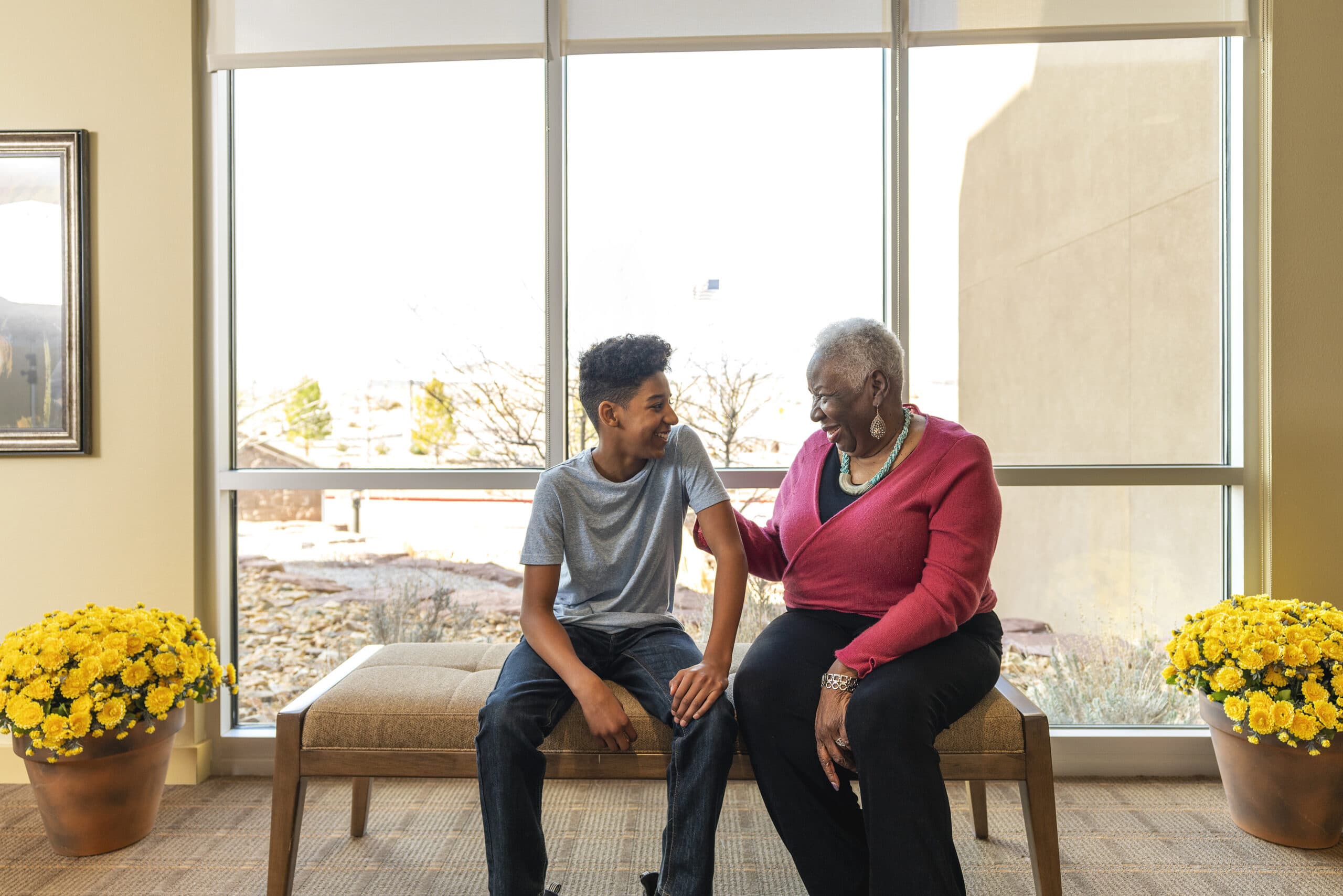 A teenage boy and an elderly woman sitting on a bench in front of a window, smiling and conversing between yellow potted flowers.