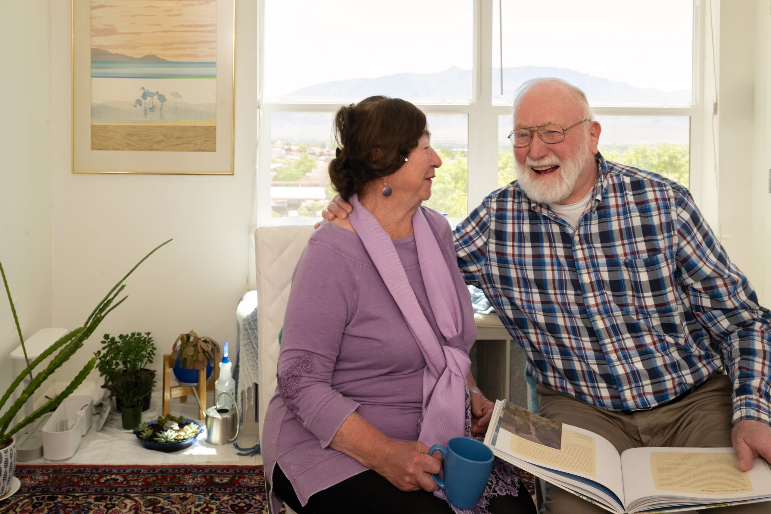 An older man and woman sitting side by side on a sofa next to a window, laughing warmly while looking at each other.