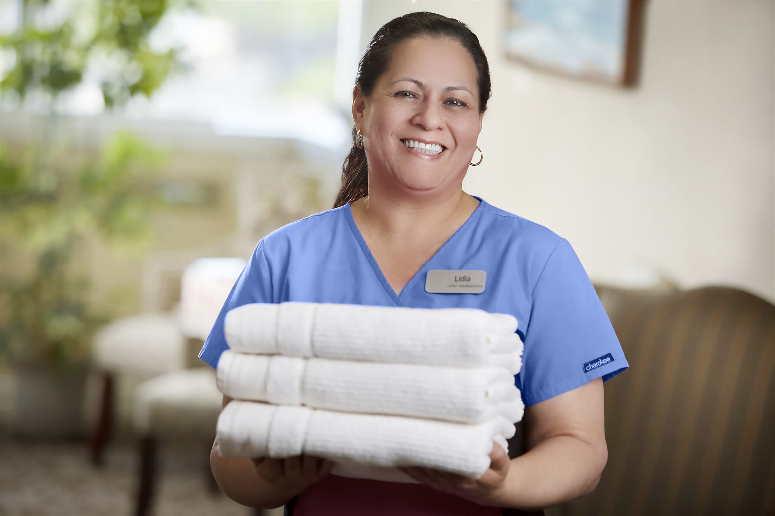 A housekeeping staff member in a blue scrub top and name badge smiling while holding a stack of folded white towels.