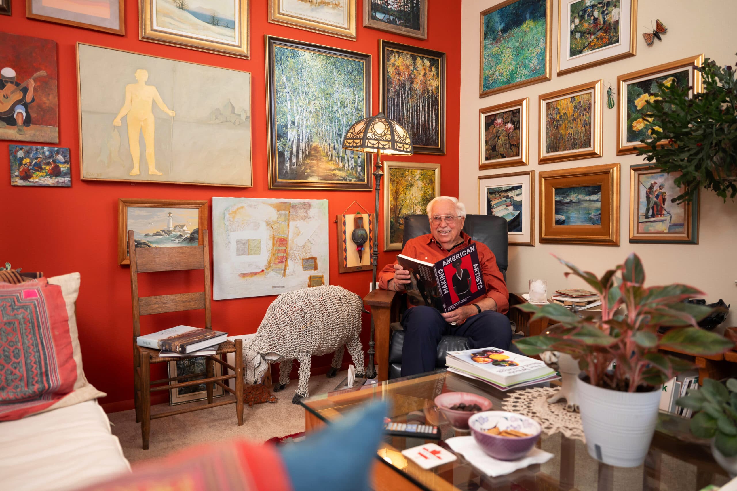 An older gentleman wearing glasses and a red shirt standing in a room filled with pottery and artwork on shelves.