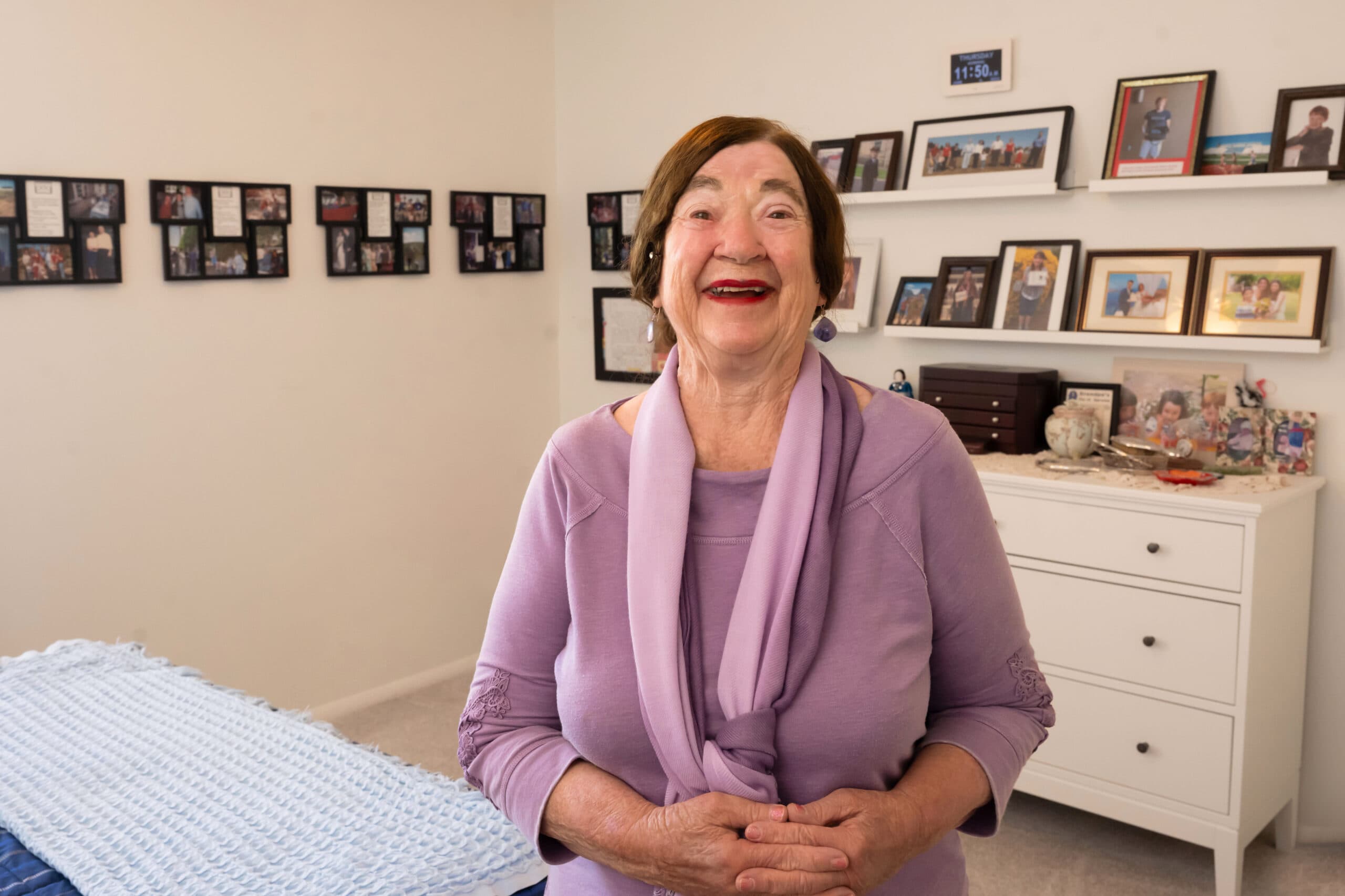 An older woman wearing a lavender top standing in a brightly lit room with family photos on shelves smiling at the camera.
