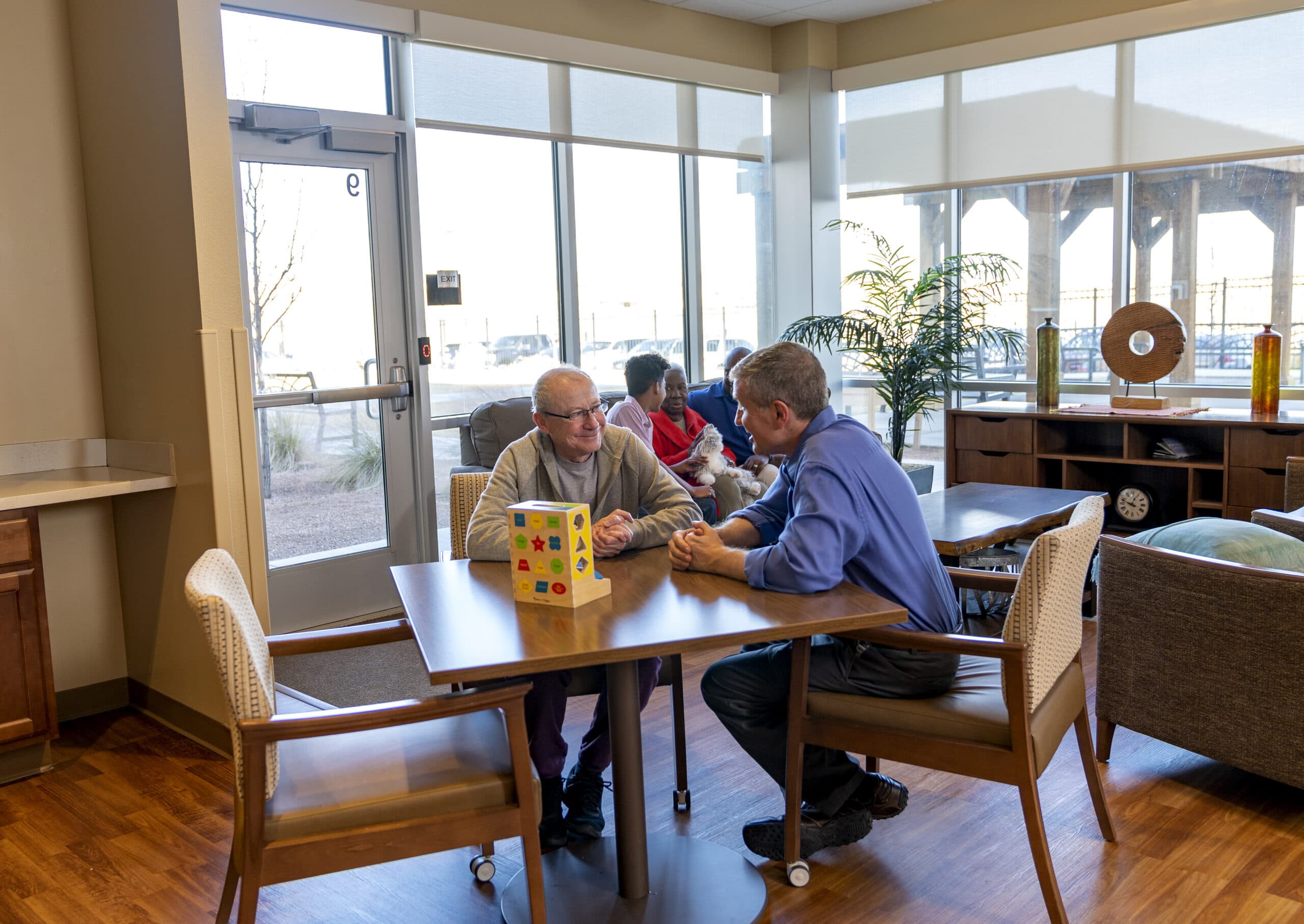 Memory care lounge with residents playing a tabletop activity near bright windows.